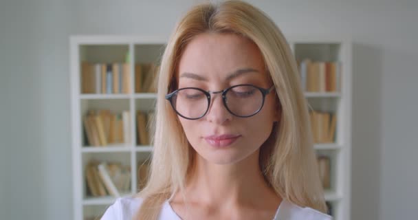 Closeup portrait of adult caucasian blonde female in glasses looking at camera smiling happily indoors in the library - Metraje, vídeo