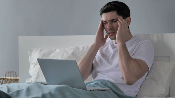 Baş ağrısı, gergin genç yatakta dizüstü bilgisayar üzerinde çalışan adam. - Fotoğraf, Görsel