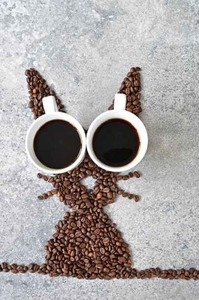 With fresh coffee beans and two cups of freshly brewed black coffee, a hare is shaped on a bright marble surface - concept with coffee beans as a gift for Easter - with room for text or other elements  - Photo, Image