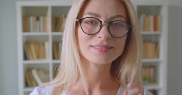 Closeup portrait of adult caucasian blonde female in glasses looking at camera smiling cheerfully indoors in the library - Metraje, vídeo