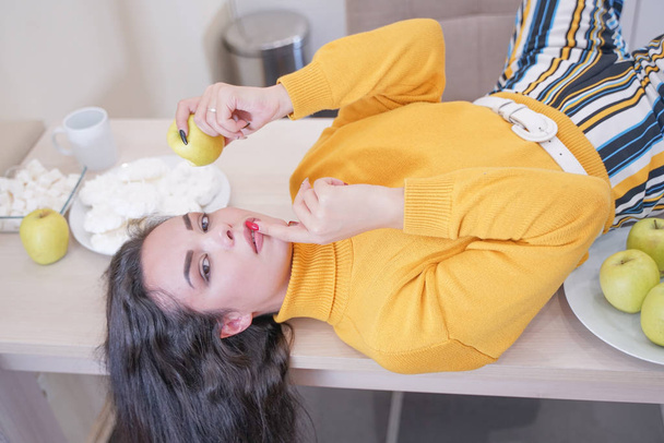 Κοντινό πορτρέτο ενός αξιαγάπητα όμορφο κορίτσι δάγκωμα ένα μήλο σε λευκή κουζίνα φόντο - Φωτογραφία, εικόνα