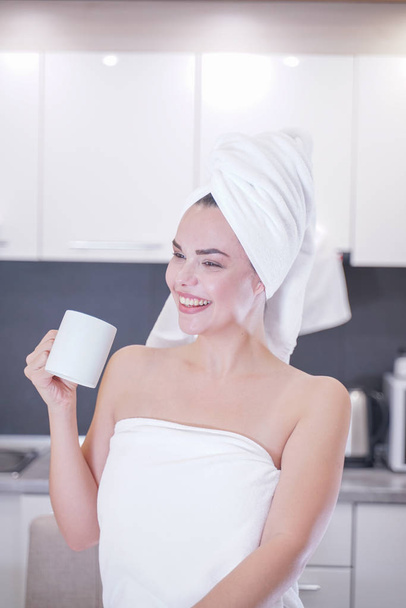 joven sentada en la cocina después de una ducha envuelta en una toalla blanca y descansando
 - Foto, Imagen