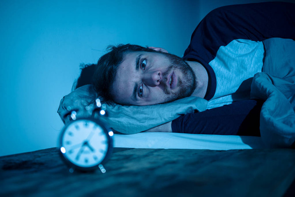 Slapeloze en wanhopige jonge Kaukasische man wakker 's nachts niet in staat om te slapen, gevoel gefrustreerd en bezorgd kijken naar klok lijden van slapeloosheid in stress en slaapstoornis concept. - Foto, afbeelding