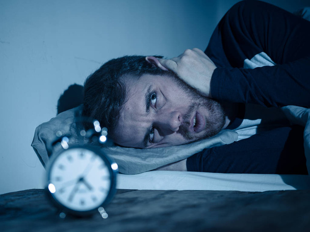 Άγρυπνος και απελπισμένος νέος Καυκάσιος άνθρωπος ξύπνιος τη νύχτα δεν είναι σε θέση να κοιμηθεί, νιώθοντας απογοητευμένος και ανήσυχος κοιτάζοντας το ρολόι που πάσχουν από αϋπνία σε άγχος και διαταραχή ύπνου αντίληψη. - Φωτογραφία, εικόνα