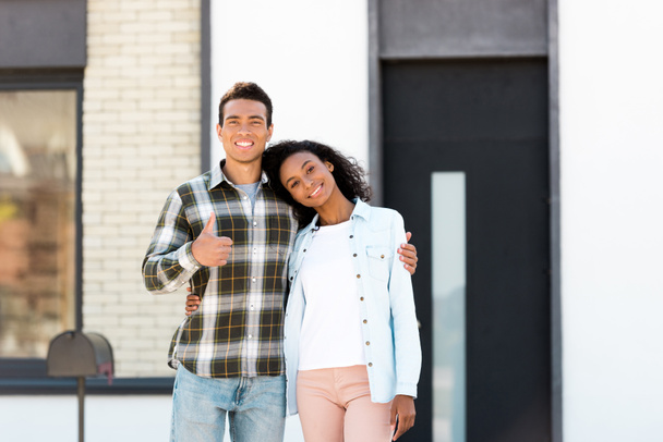 african american husband and wife standing near new house and looking at camera while man showing thumb up  - Фото, зображення