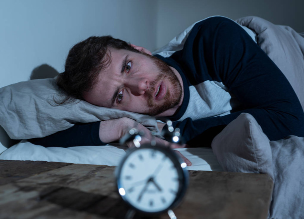 Insomnia Stress and Sleeping disorder concept. Sleepless desperate young caucasian man awake at night not able to sleep, feeling frustrated and worried looking stressed and concerned at alarm clock. - Фото, изображение