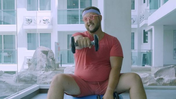 Смешной толстый мужчина в розовых очках и в розовой футболке обручен с гантелями на мяче в спортзале, изображающем девушку
 - Фото, изображение
