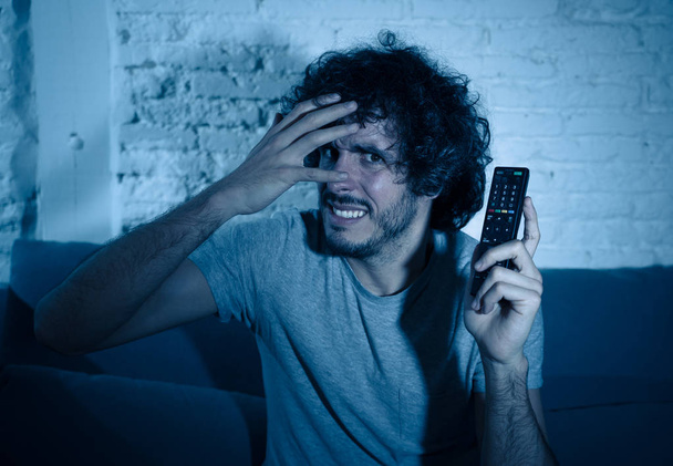 Lifestyle portrait of young man feeling scared and shocked making fear, anxiety gestures while watching television holding remote control. In horror and violence on TV and internet and mass media. - Photo, Image
