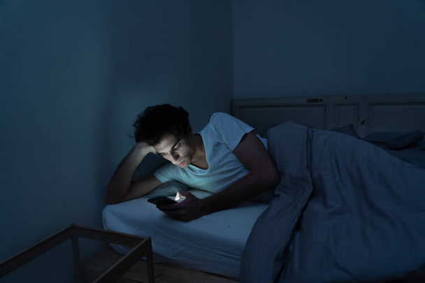 Addicted man chatting and surfing on the Internet with smart phone late at night in bed. Bored, sleepless and tired in dark room with moody light. In insomnia and mobile addiction concept. - Fotografie, Obrázek