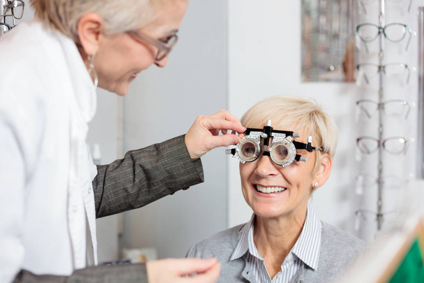 Χαμογελαστή ώριμη γυναίκα που έχει οφθαλμολογική εξέταση και μέτρηση διοπτέρ από μια θηλυκή οπτομέτρη στην οφθαλμολογική κλινική - Φωτογραφία, εικόνα