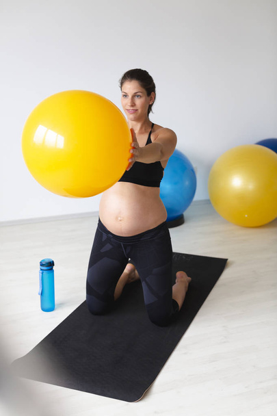 beautiful young pregnant woman in black sportswear holding aerobic ball in raised arms and standing on knees on mat while doing fitness exercise in light studio - Fotó, kép