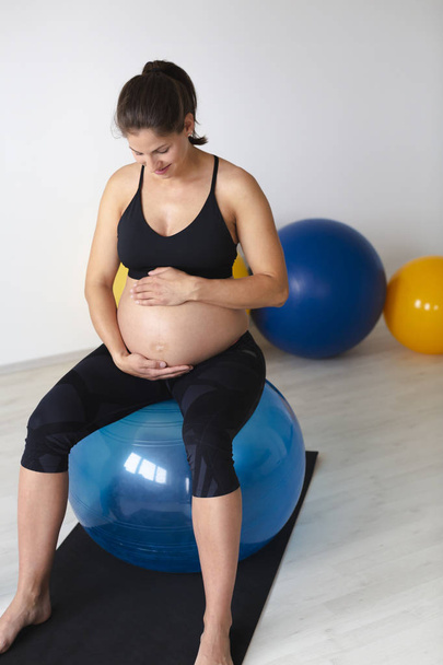 όμορφη νεαρή έγκυος γυναίκα σε μαύρο αθλητικά ρούχα αγκαλιάζει απαλά κοιλιά με τα χέρια, ενώ κάθονται στο γυμναστήριο μπάλα και χαλαρωτικό μετά την άσκηση γυμναστικής σε φωτεινό στούντιο - Φωτογραφία, εικόνα