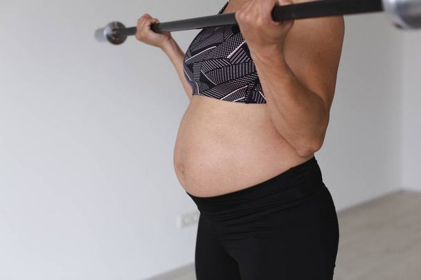beautiful young pregnant woman in black sportswear doing fitness exercise with barbell bar in light studio - 写真・画像