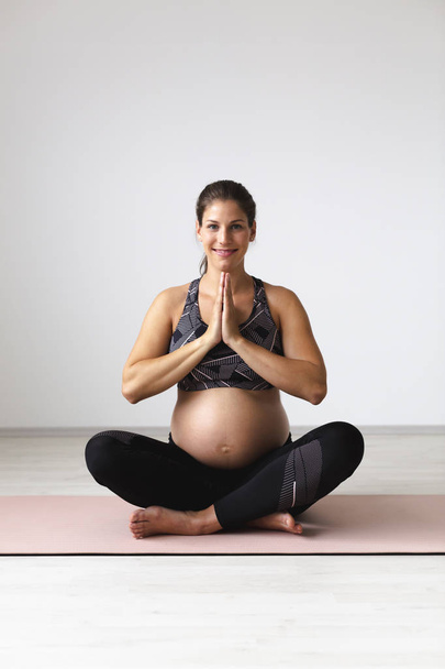 beautiful young pregnant woman in black sportswear holding hands in namaste while practising yoga and meditating in light studio - Foto, Imagem