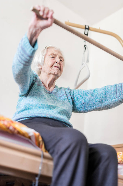 Elderly 96 years old woman exercising with a stick sitting on her bad. - Photo, Image