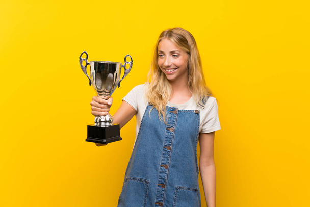 Blonde young woman over isolated yellow background holding a trophy - Fotografie, Obrázek
