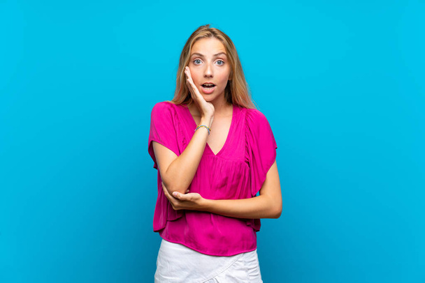 Blonde jonge vrouw over geïsoleerde blauwe achtergrond verrast en geschokt terwijl op zoek naar rechts - Foto, afbeelding