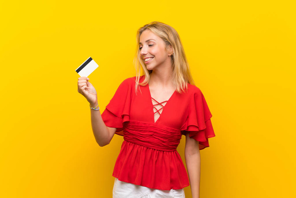Blonde young woman over isolated yellow background holding a credit card - Photo, Image