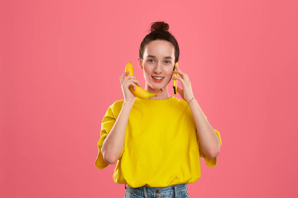 Χαμογελαστή μοντέλο με ένα κουλούρι μαλλιών σε ένα κίτρινο t-shirt κρατώντας μπανάνα και κίτρινο smartphone - Φωτογραφία, εικόνα