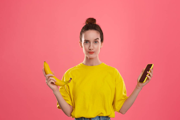 Model with a hair bun in a yellow t-shirt holding banana and yellow smartphone - Foto, afbeelding