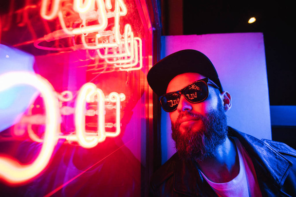 Hipster handsome man on the city streets being illuminated by neon signs. He is wearing leather biker jacket or asymmetric zip jacket with black cap, jeans and sunglasses. - Фото, зображення