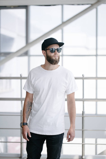 Hipster handsome male model with beard wearing white blank t-shirt and a baseball cap with space for your logo or design in casual urban style - Valokuva, kuva