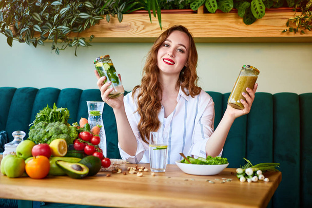 junge Frau trinkt Smoothie und frisches Wasser im schönen Interieur mit grünen Blumen im Hintergrund und frischem Obst und Gemüse auf dem Tisch. Konzept für gesunde Ernährung. Vegane Mahlzeit und Detox-Menü - Foto, Bild