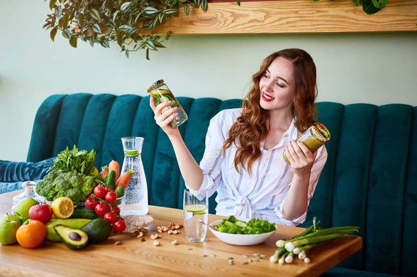 junge Frau trinkt Smoothie und frisches Wasser im schönen Interieur mit grünen Blumen im Hintergrund und frischem Obst und Gemüse auf dem Tisch. Konzept für gesunde Ernährung. Vegane Mahlzeit und Detox-Menü - Foto, Bild