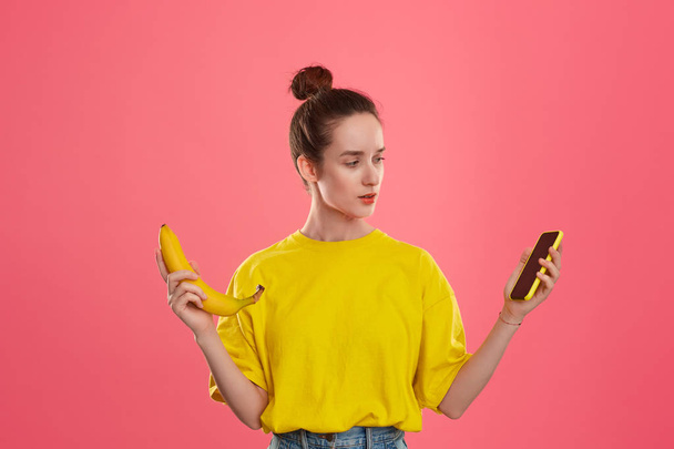 Μοντέλο με ένα κουλούρι μαλλιών σε ένα κίτρινο t-shirt κρατώντας μπανάνα και κίτρινο smartphone - Φωτογραφία, εικόνα