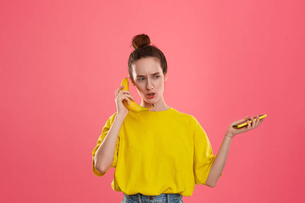 Modelo confuso con un moño de pelo en una camiseta amarilla sosteniendo plátano hablando en un teléfono inteligente amarillo
  - Foto, imagen