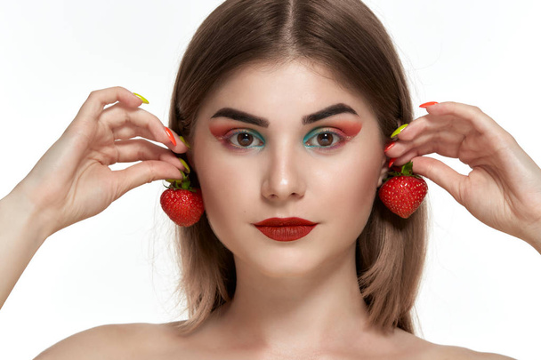 Close-up portret van een mooie jonge vrouw met felle kleur make-up holding aardbei in de buurt van het gezicht. - Foto, afbeelding