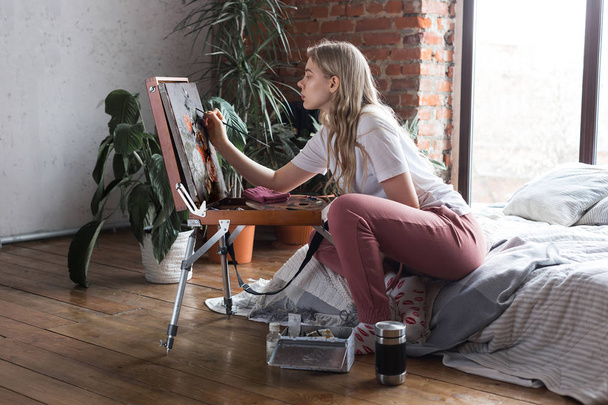 ブラシとパレットを持つ若いかわいい女の子は、イーゼル描画絵の近くに座っています。アート、創造性、趣味、描画プロセス. - 写真・画像