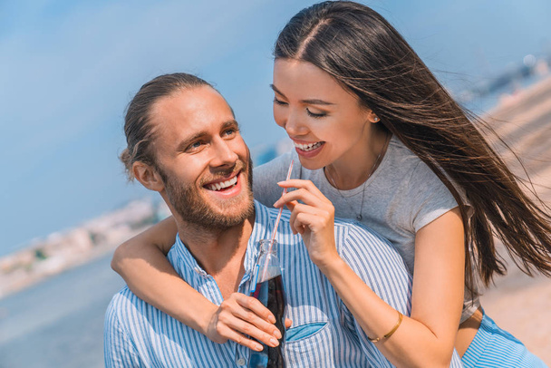 Happy caucasian couple enjoying themselves outdoors. Handsome young guy piggybacking his girlfriend drinking soft drink outdoors. - Foto, Imagen