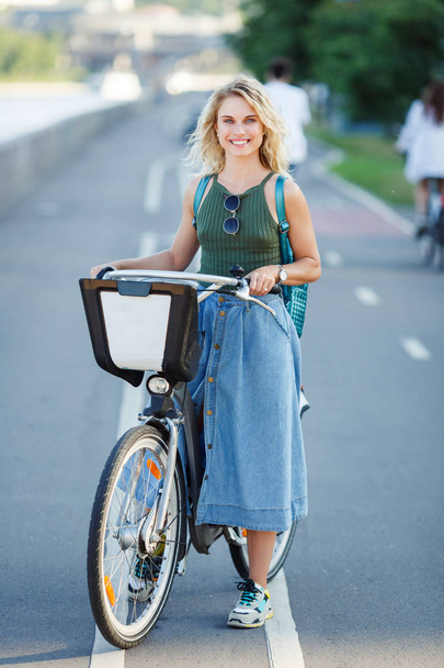 Photo of young blonde in long denim skirt standing next to bike on road in city - Foto, Imagen