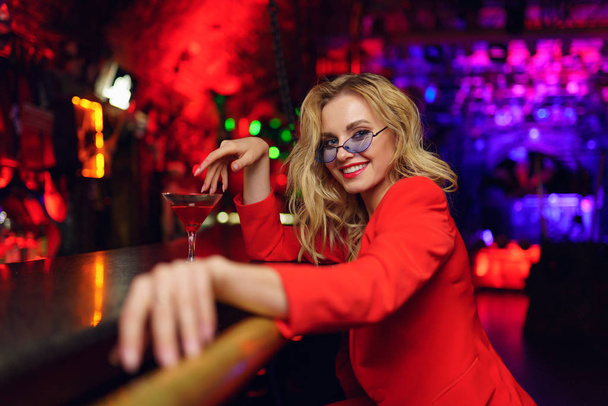 Zdjęcie Happy Blonde z okularami i czerwonym garniturze siedząc w barze z kieliszkami koktajlową w klubie nocnym - Zdjęcie, obraz