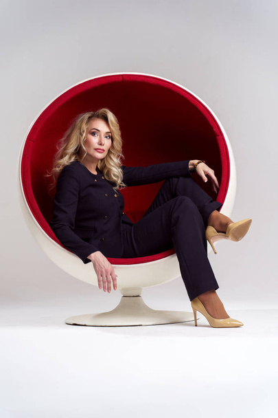 Image of blonde woman with long curly hair in black suit sitting in round chair isolated on white background - Photo, Image