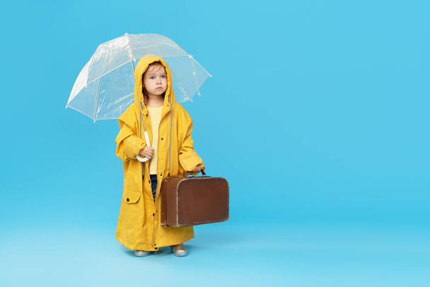 Happy funny child with transparent umbrella posing on blue studio background. Girl is wearing yellow raincoat and rubber boots. Holds a vintage travel suitcase - Photo, image