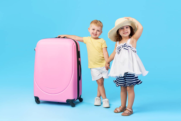 Happy kids sitting on colorful pink suitcase prepared for summer vacation. Young travelers. Little girl and boy, sister and brother, having fun isolated on blue background - Фото, изображение
