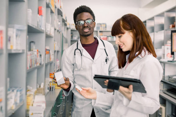 Two pharmacist looking for medicinal drug. Male African pharmacist holding a medicine with female standing by and using digital tablet in pharmacy. - 写真・画像