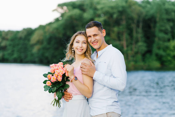 a loving couple a guy and a girl are standing on the Bank of a river or lake. first date. the girl is holding a bouquet of roses, the guy hugs the girl, they smile and - Photo, Image