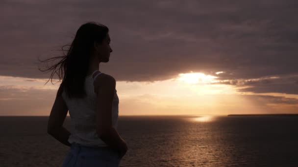 Sympatyczny brunetka dziewczyna stoi i patrząc na zachód słońca nad Morzem Czarnym w SLO-mo arty widok z uroczej Brunetki dziewczyna z latające długie luźne włosy w dżinsy i Koszulka bez rękawów uzyskiwanie inspiracji na fale Morza Czarnego w Nicei zachód słońca w SLO-mo - Materiał filmowy, wideo