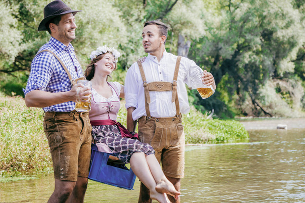happy young friends in Bavarian costumes holding glasses of beer and walking outdoor  - Foto, Imagem