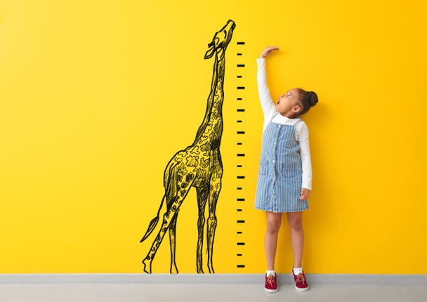 Удивлённая афроамериканка, измеряющая высоту рядом с цветной стеной с нарисованным жирафом
 - Фото, изображение