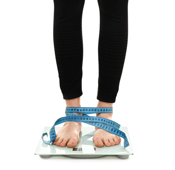 Legs of young woman with measuring tape standing on scales against white background. Weight loss concept - 写真・画像
