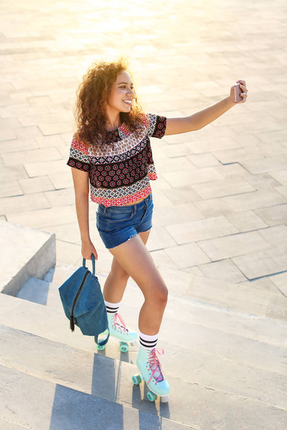 Beautiful young woman on roller skates taking selfie outdoors - Фото, изображение
