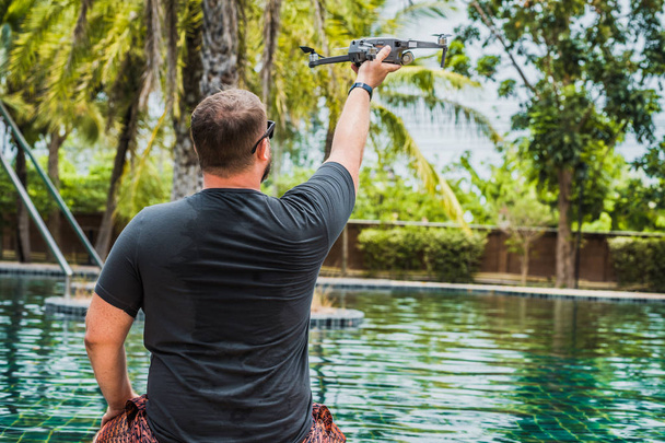 Un hombre simpático lanza un dron para el vuelo, con el que se pueden tomar fotos y videos, cerca de la piscina, ver hacia atrás
. - Foto, Imagen