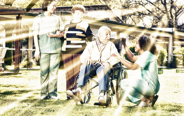 Two female doctors helping elderly man and woman with physical disability ih rehab facility garden, with sun rays. - Photo, Image