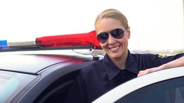 Policewoman in sunglasses smiling, looking into camera, standing near patrol car - Фото, изображение