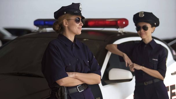 Positive police women standing near car, talking and smiling, break from work - 写真・画像