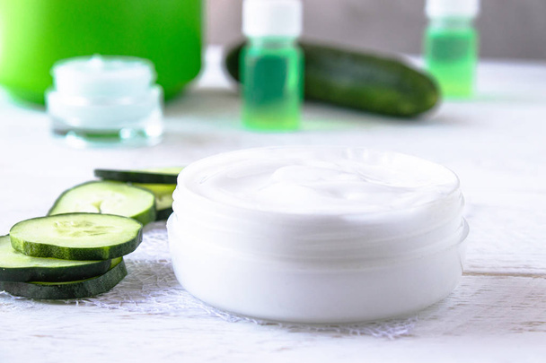 Face cream in a white jar with cucumber extract next to slices of fresh cucumbers on a white wooden background. - Foto, afbeelding